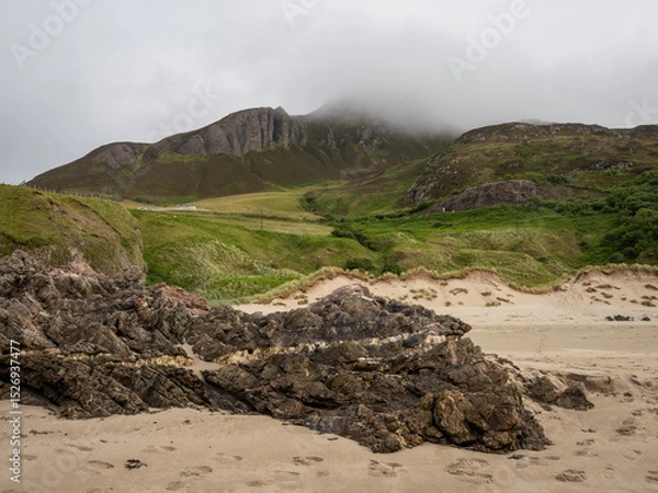 Fototapeta Sandy beach with rocks in the foreground and mountain peak shrouded in mist in the background
