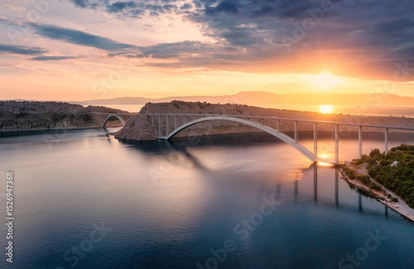Obraz Aerial view of bridge and blue sea at colorful sunset. Modern Krk bridge island during golden hour, mountains, rocks, reflection in water and orange sky. Top drone view of bridges. Architecture