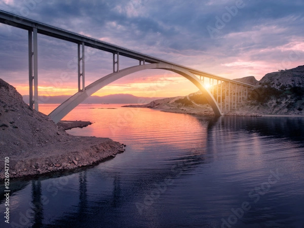 Obraz Aerial view of bridge and blue sea at colorful sunset. Modern Krk bridge island during golden hour, mountains, rocks, reflection in water and cloudy purple sky. Top drone view of bridges. Architecture