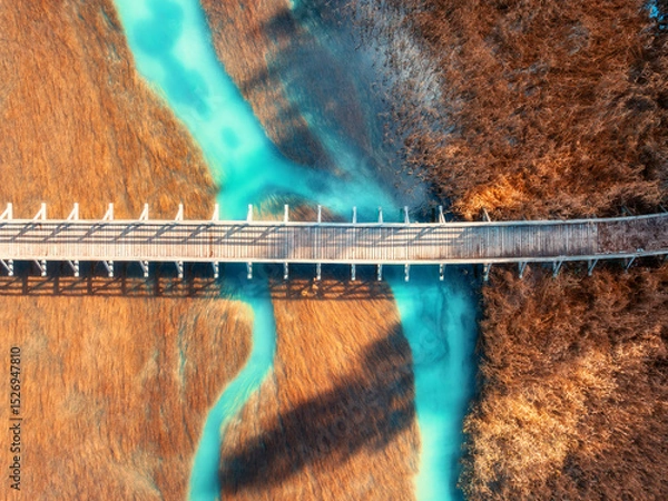 Obraz Aerial view of wooden bridge, azure river and orange grass at sunset in golden autumn. Zelenci lake, Slovenia, Triglav national park in fall. Top drone view of bridge, green water. Nature. Landscape