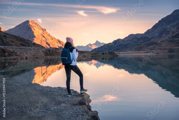 Obraz Young woman hiker with backpack standing on the shore of a mountain lake enjoying the sunrise over the swiss alps in autumn. Concept of adventure, travel, healthy lifestyle. Girl on the lake in fall