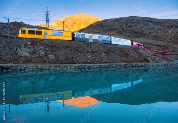 Obraz Bernina express train passing by Bianco lake with colorful reflections of the train and mountains during sunset in the swiss alps in autumn. Fall in Switzerland. Yellow modern train with red wagons