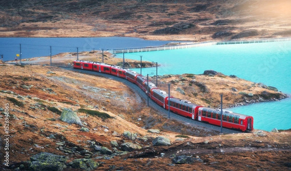 Obraz Aerial view of modern red train near azure mountain lake and dramatic cloudy sky at sunset in autumn. Bernina Express, Switzerland. Top view of train in Alps, winding railroad, orange hills in fall
