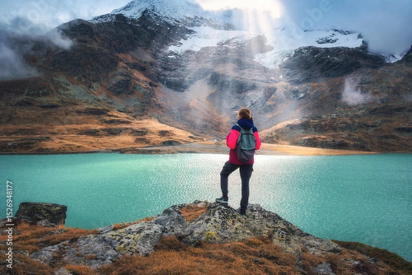 Obraz Young woman hiker with backpack standing on a rock with view on turquoise alpine lake, sunbeams, snowy mountain and glacier in the background in fall. Girl on mountain trail in Swiss Alps in autumn