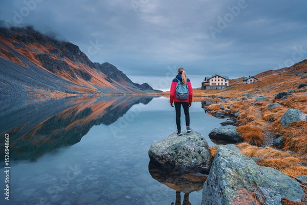 Obraz Young woman hiker with backpack on a rock in autumn. Girl and breathtaking view of a mountain alpine lake reflecting the surrounding peaks, orange grass and cloudy dramatic sky. Switzerland in fall