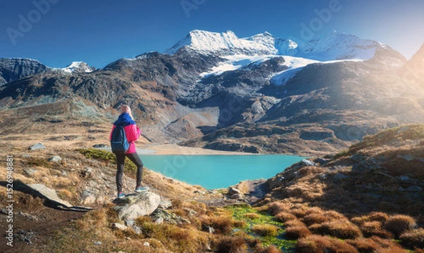 Obraz Young woman hiker with backpack standing on a rock admiring a beautiful turquoise alpine lake with snowy mountain and glacier in the background in fall. Girl on mountain trail in Swiss Alps in autumn