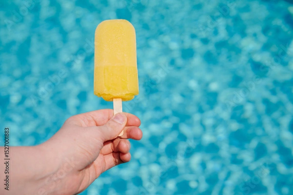 Fototapeta Bright yellow popsicle held above shimmering blue pool water on a warm summer day