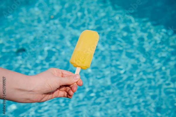 Fototapeta Bright yellow popsicle held above a shimmering blue swimming pool during a sunny summer day