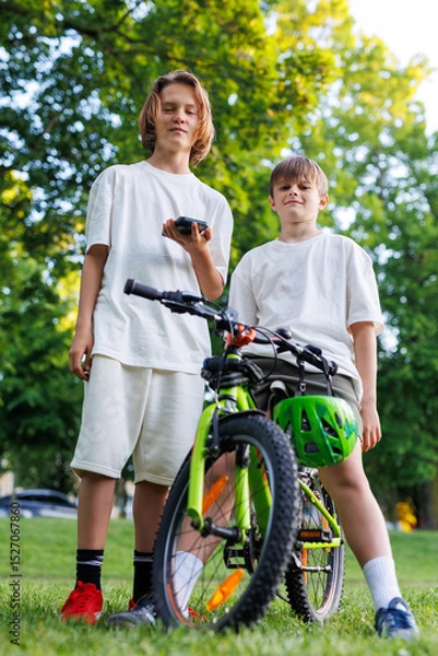 Fototapeta Two boys in white T-shirts pose confidently outdoors with a green mountain bike. One holds a phone, the other rests on the handlebar, both looking down with cocky expressions in summer park setting