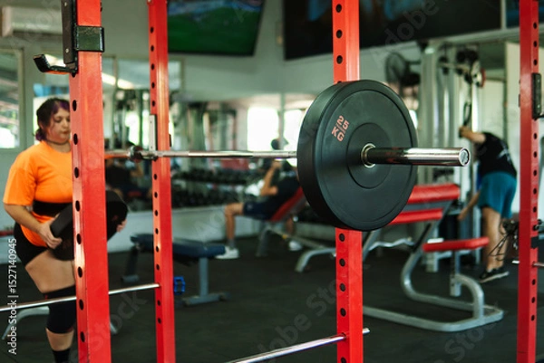 Fototapeta View of an overweight young Latina woman carrying weights and preparing to workout at a gym. Pushing herself. Improvement and well-being.