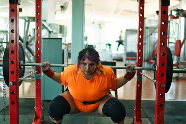 Fototapeta View of a beautiful, overweight young Latina lifting weights and working out at a gym. Positively, she's pushing herself to the limit. She's improving and feeling better.