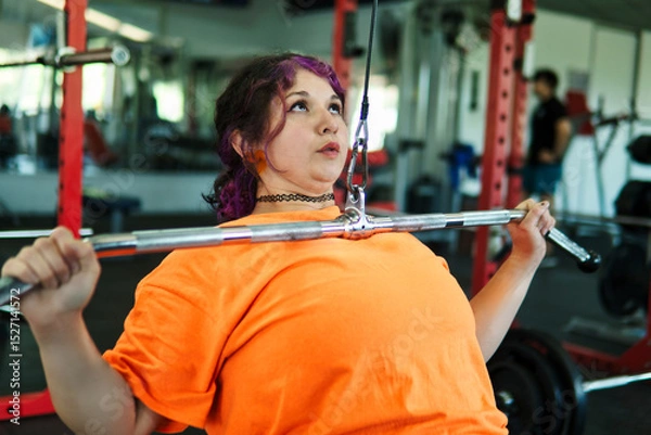 Fototapeta Beautiful, overweight Latina woman concentrating on lifting weights on an exercise machine at a gym. Wearing colorful clothes, hair, and retro makeup, she's positive. She's pushing herself.