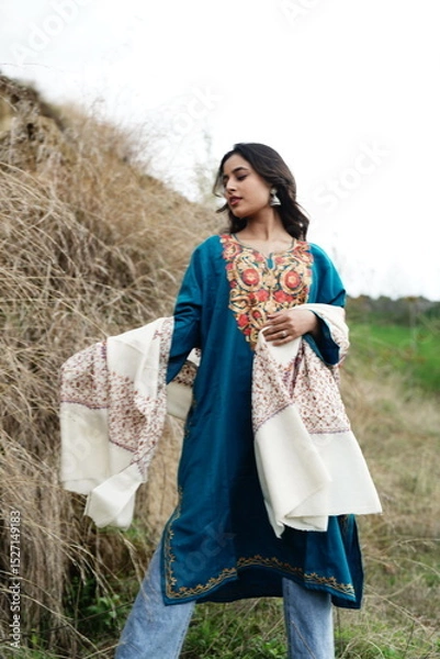 Fototapeta A female Kashmiri model wearing a traditional pheran and shawl, posing for the artisanal brand Kaar Kash, with a richly embroidered curtain as a backdrop in a serene outdoor setting.

