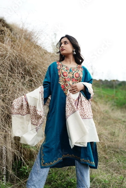 Fototapeta A female Kashmiri model wearing a traditional pheran and shawl, posing for the artisanal brand Kaar Kash, with a richly embroidered curtain as a backdrop in a serene outdoor setting.


