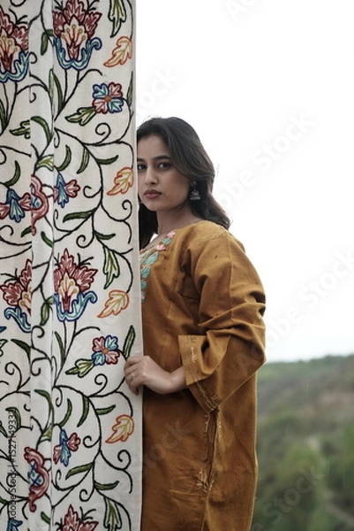 Fototapeta A female Kashmiri model wearing a traditional pheran and shawl, posing for the artisanal brand Kaar Kash, with a richly embroidered curtain as a backdrop in a serene outdoor setting.

