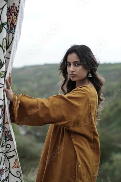 Fototapeta A female Kashmiri model wearing a traditional pheran and shawl, posing for the artisanal brand Kaar Kash, with a richly embroidered curtain as a backdrop in a serene outdoor setting.

