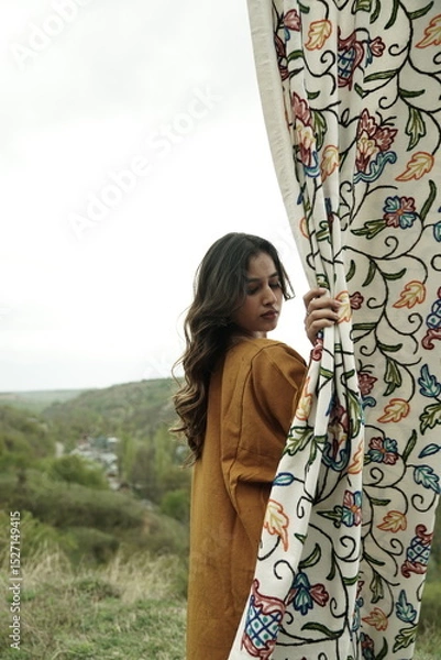 Fototapeta A female Kashmiri model wearing a traditional pheran and shawl, posing for the artisanal brand Kaar Kash, with a richly embroidered curtain as a backdrop in a serene outdoor setting.

