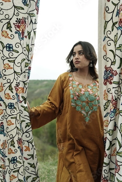 Fototapeta A female Kashmiri model wearing a traditional pheran and shawl, posing for the artisanal brand Kaar Kash, with a richly embroidered curtain as a backdrop in a serene outdoor setting.

