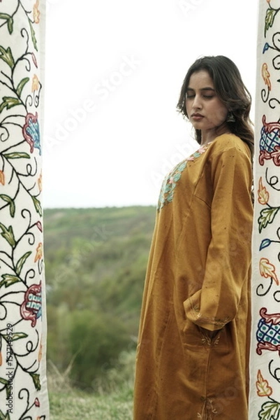 Fototapeta A female Kashmiri model wearing a traditional pheran and shawl, posing for the artisanal brand Kaar Kash, with a richly embroidered curtain as a backdrop in a serene outdoor setting.

