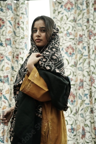 Fototapeta A female Kashmiri model wearing a traditional pheran and shawl, posing for the artisanal brand Kaar Kash, with a richly embroidered curtain as a backdrop in a serene outdoor setting.

