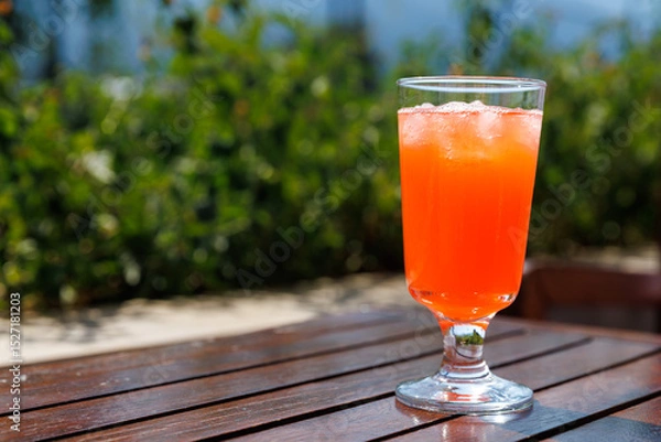 Fototapeta Bright orange drink served in glass with ice cubes on wooden table outdoor with blurred green foliage in background, summer concept, copy space.