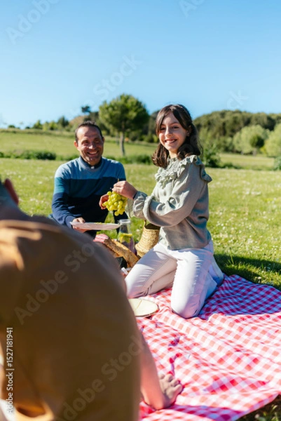 Fototapeta Family having fun during a picnic on a sunny day, eating grapes and other food, enjoying togetherness and nature