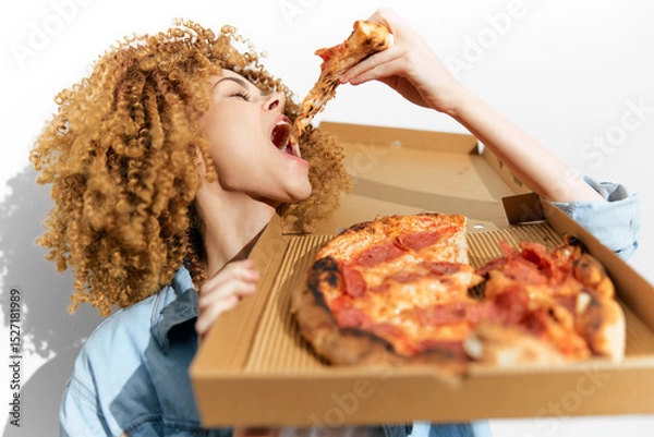 Fototapeta A joyful young woman enthusiastically enjoying pizza from a delivery box, showcasing a fun and vibrant atmosphere with her curly hair and casual outfit