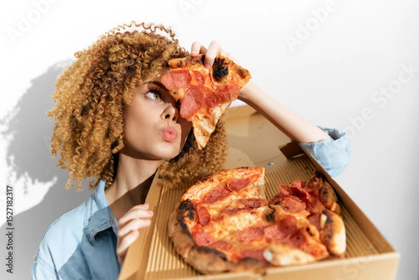 Fototapeta A young woman with curly hair playfully poses with a pizza box, holding a slice of pepperoni pizza and enjoying the moment, showcasing fun, food, and joy