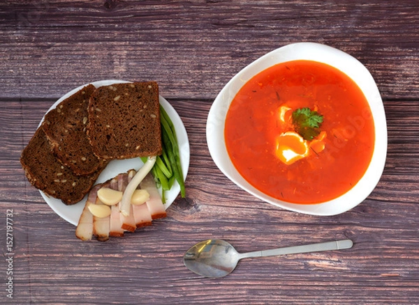 Obraz Borscht with sour cream in a deep plate, spoon and dish with rye bread, garlic, lard and green onions on a wooden table.