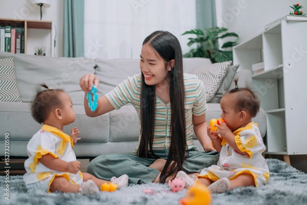 Obraz Portrait of Young Asian mother shares a happy moment with her twin daughters while sitting on a soft carpet in a cozy room at home. Leisure activities with family
