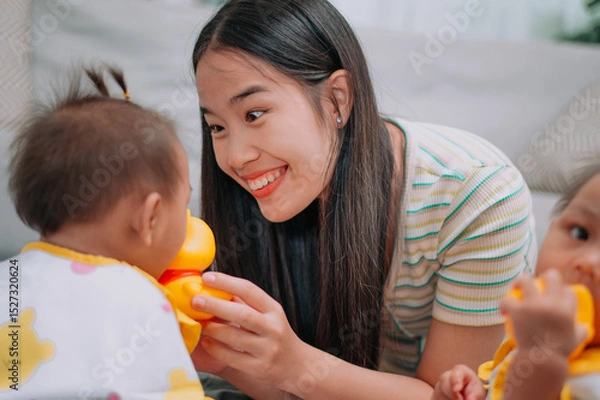Obraz Selective focus shot, Young Asian mother shares a happy moment with her twin daughters while sitting on a soft carpet in a cozy room at home. Leisure activities with family