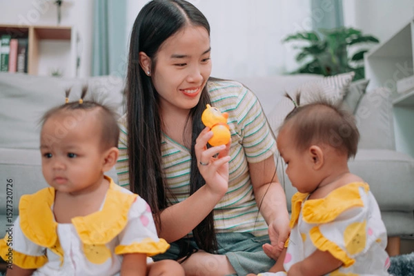Obraz Portrait of Young Asian mother shares a happy moment with her twin daughters while sitting on a soft carpet in a cozy room at home. Leisure activities with family