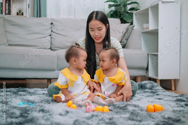 Obraz Portrait of Young Asian mother shares a happy moment with her twin daughters while sitting on a soft carpet in a cozy room at home. Leisure activities with family
