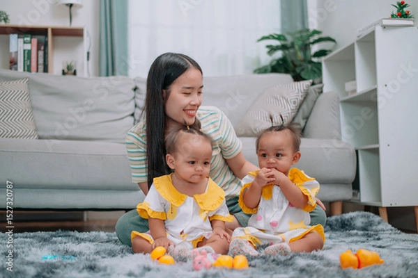 Obraz Portrait of Young Asian mother shares a happy moment with her twin daughters while sitting on a soft carpet in a cozy room at home. Leisure activities with family