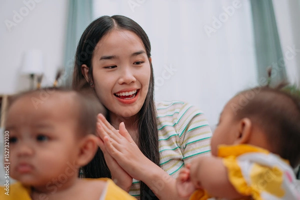 Obraz Selective focus shot, Young Asian mother shares a happy moment with her twin daughters while sitting on a soft carpet in a cozy room at home. Leisure activities with family
