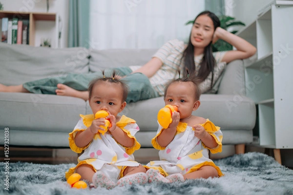 Obraz Portrait of Asian mother lies on the couch and takes care of her twins while they play with toys in the living room at home.
