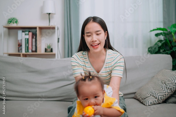Obraz Portrait of Asian mother sits on the sofa holding her little daughter and playing with her happily in the living room at home.