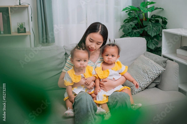 Obraz High angle view shot, A young mother sits on a sofa and gently holds her twin daughters in living room at home.