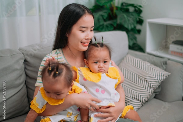 Obraz Portrait of Young mother sits on a sofa and gently holds her twin daughters in living room at home.