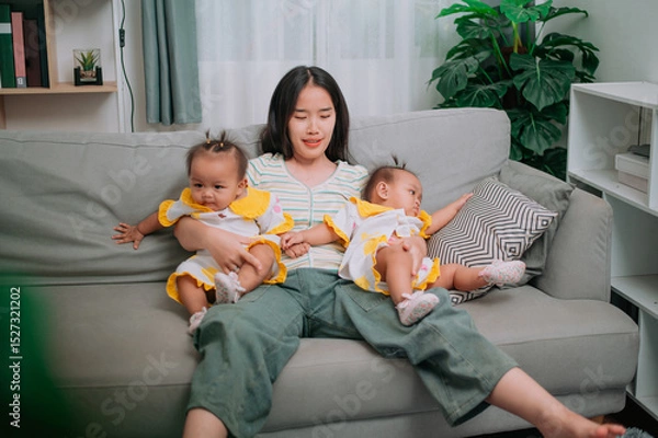 Obraz Portrait of Young mother sits on a sofa and gently holds her twin daughters in living room at home.