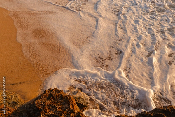 Fototapeta Waves wash the sandy beach in the setting sun view from above. The texture of sand and foamy waves. Beautiful natural background