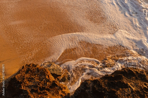 Fototapeta Waves wash the sandy beach in the setting sun view from above. The texture of sand and foamy waves. Beautiful natural background