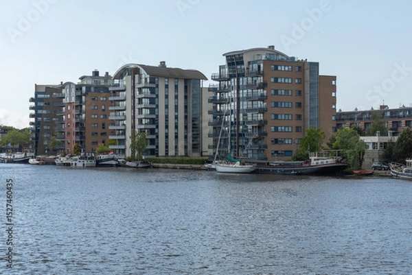 Obraz This contemporary waterside scene in Wormerveer, Netherlands, showcases modern residential buildings with unique architecture and boats docked along the calm water, under a bright sky.