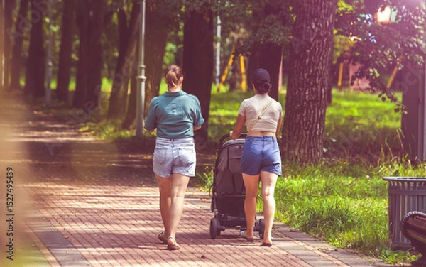 Fototapeta Two girls with a baby stroller are walking in the summer park
