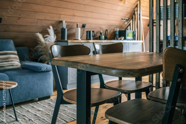 Obraz Close-up of modern wooden dining table and chairs in cozy Scandinavian style kitchen with wooden walls, blue sofa, and kitchen countertop
