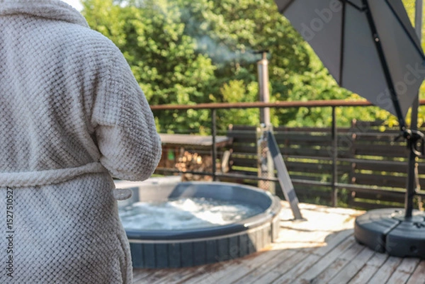 Obraz Person in white bathrobe standing near outdoor hot tub with bubbling water on wooden terrace, surrounded by green trees and backyard privacy fence
