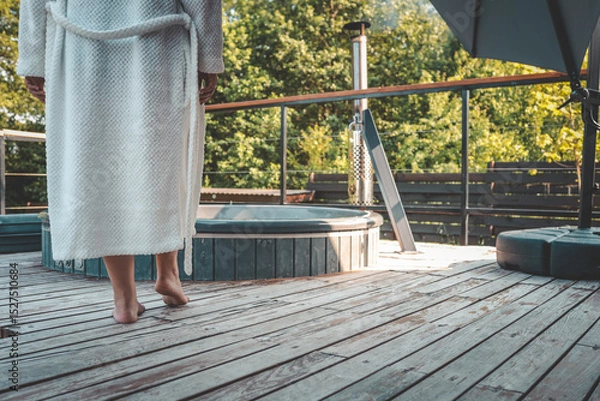 Obraz Close-up of barefoot person in white bathrobe walking on wooden terrace towards outdoor hot tub on a sunny day, relaxation and spa concept