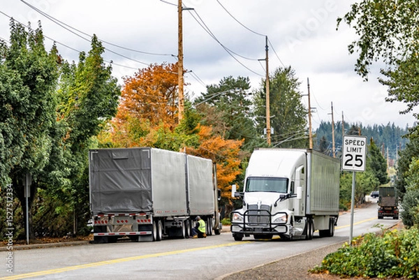 Obraz Truck driver of the big rig semi truck with two conestoga semi trailers repairs a wheel on the road putting himself at risk while an oncoming semi truck driving on the autumn local road