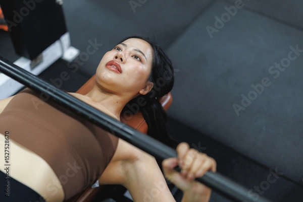 Fototapeta Determined asian woman lifting weights while bench pressing in gym