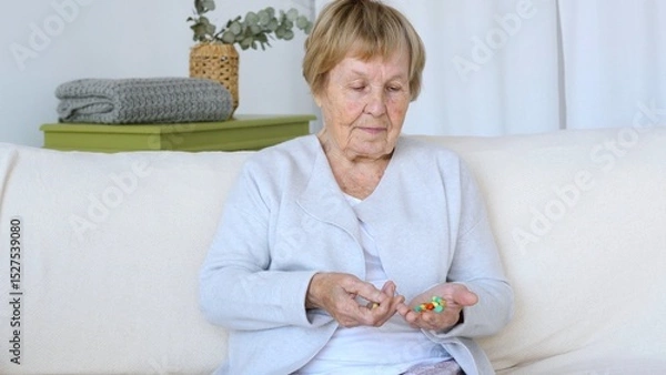 Fototapeta Senior woman sitting on a comfortable sofa, carefully examining variety of pills in her hands, showcasing the challenges of managing medication in old age